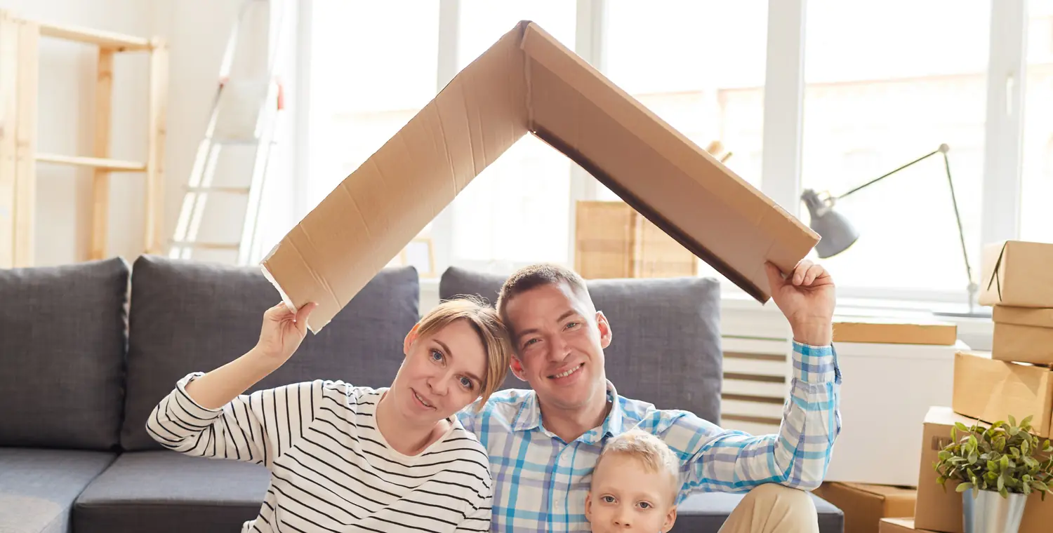 Une famille assise au milieu des cartons d'un emménagement, un toit de carton tenu à bout de bras, en tête du guide « Hypothèque non-résident »